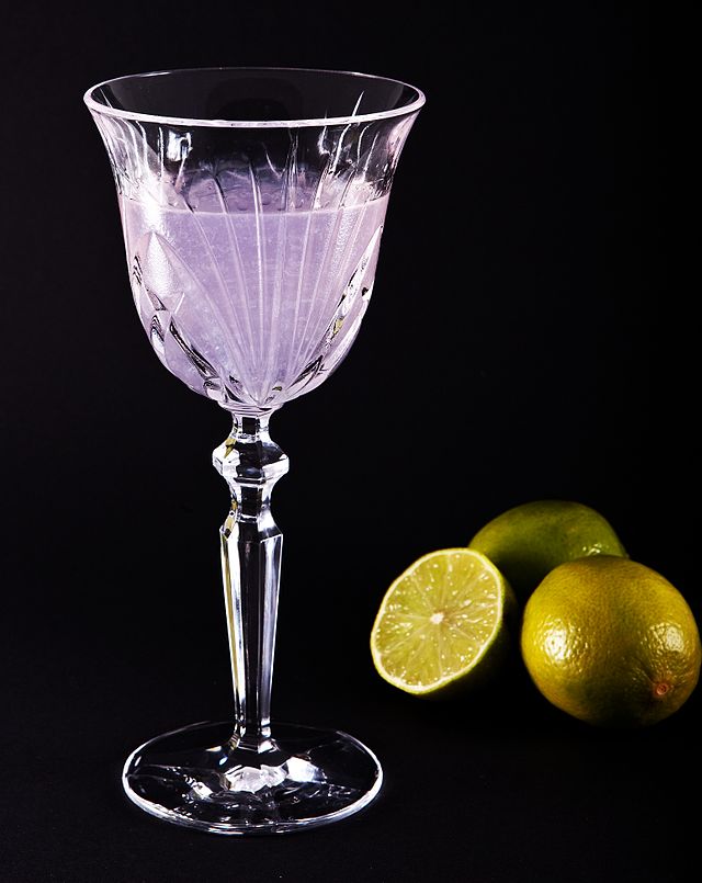 An Aviation Cocktail in a fancy looking glass with limes behind it.