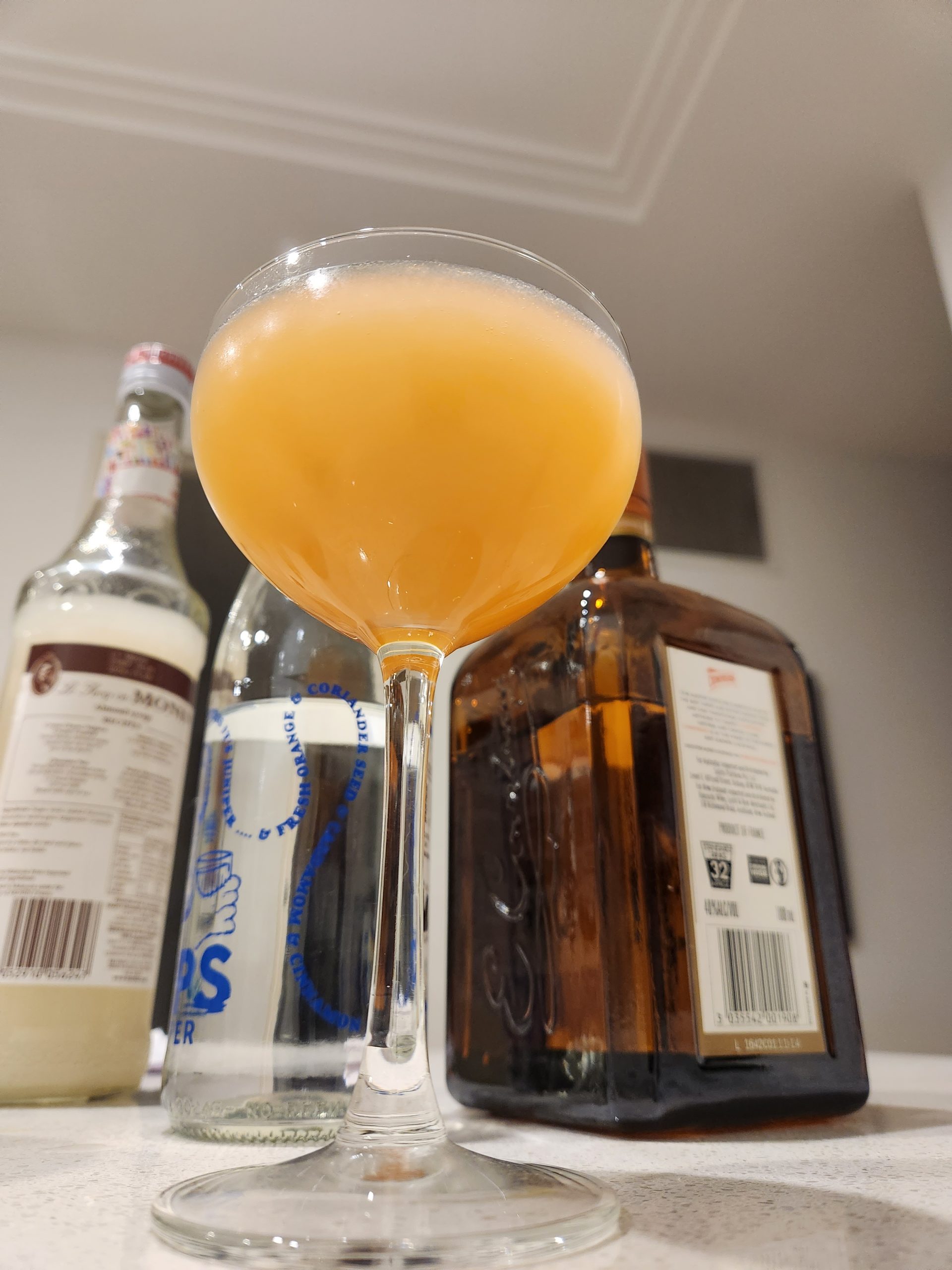 A Bronx cocktail in a coupe glass with various ingredients behind it.