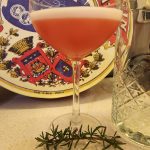A Winter Sour cocktail in a coupe glass with a sprig of rosemary in front of it.